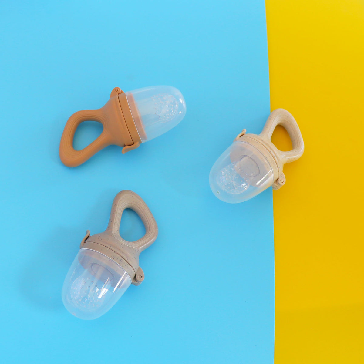 Three capped baby food feeders with peach, cream and gray handles on a blue and yellow background